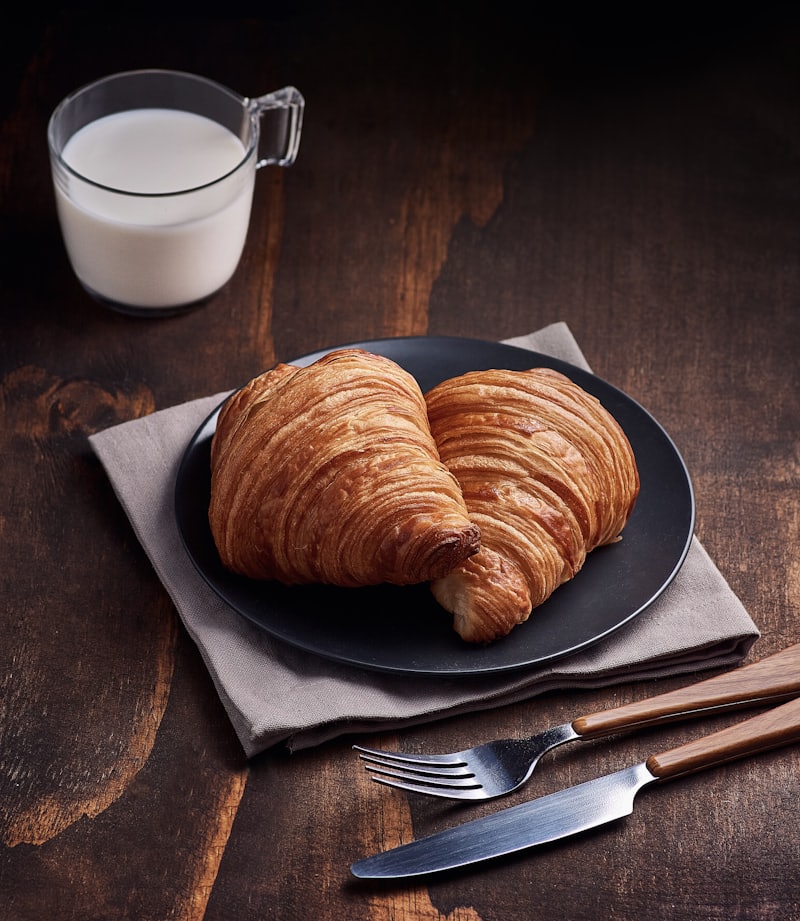 Two golden croissants on a dark plate beside a glass of milk