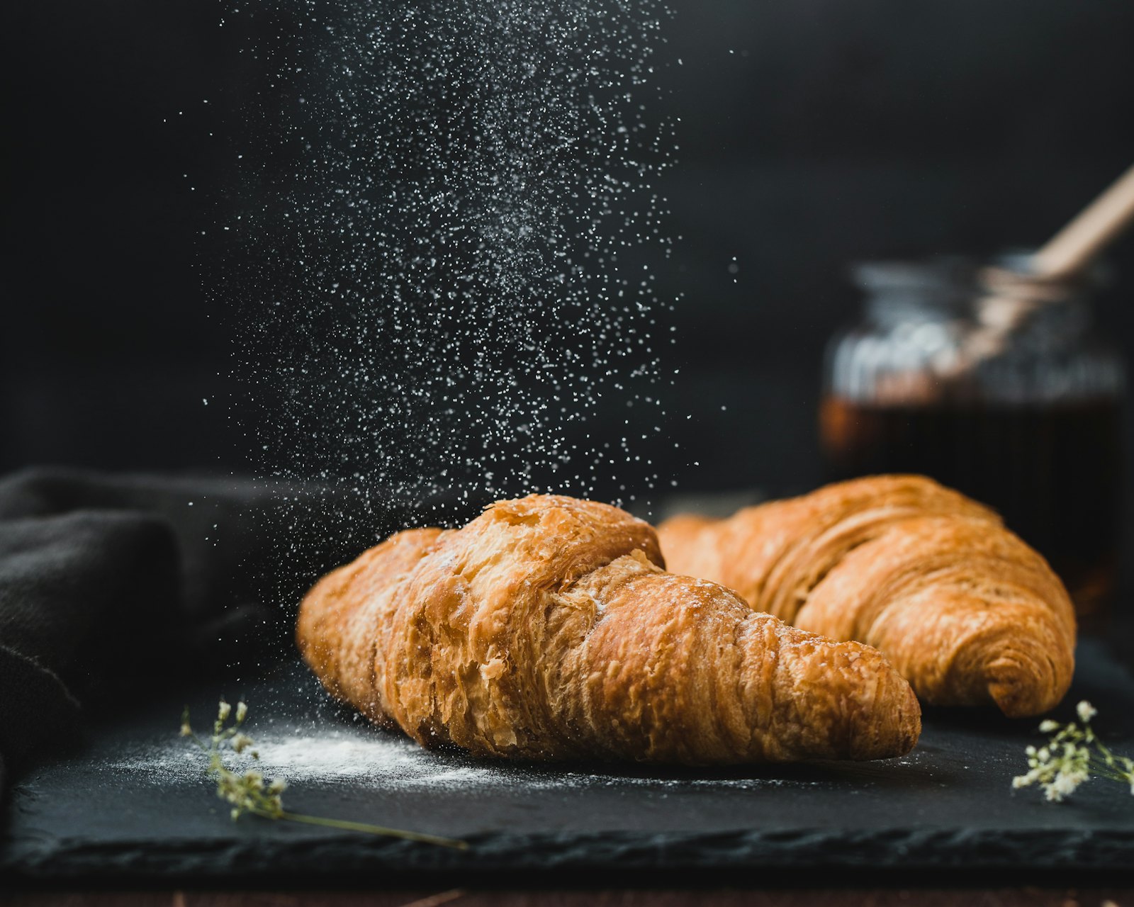 A golden butter croissant dusted with a cloud of icing sugar on a dark wooden board